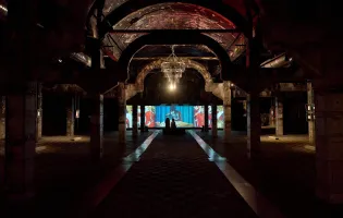 Nave industrial a oscuras con una gran lámpara iluminada y pantallas al fondo