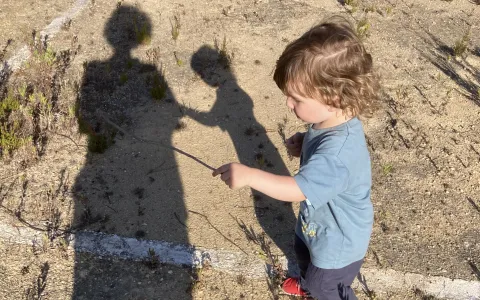 Un niño de 2 años juega con un palo observado por su madre. las sombras de ambos se proyectan en el suelo