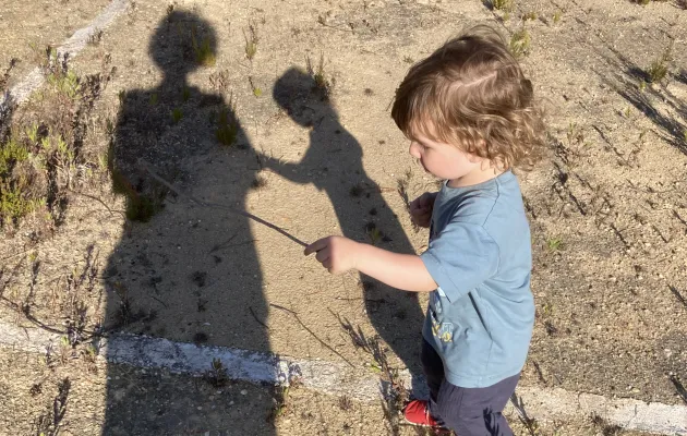 Un niño de 2 años juega con un palo observado por su madre. las sombras de ambos se proyectan en el suelo