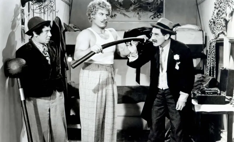 Hombre con bigote y sobrero, Groucho Marx, mira incrédulo a un forzudo que dobla una barra