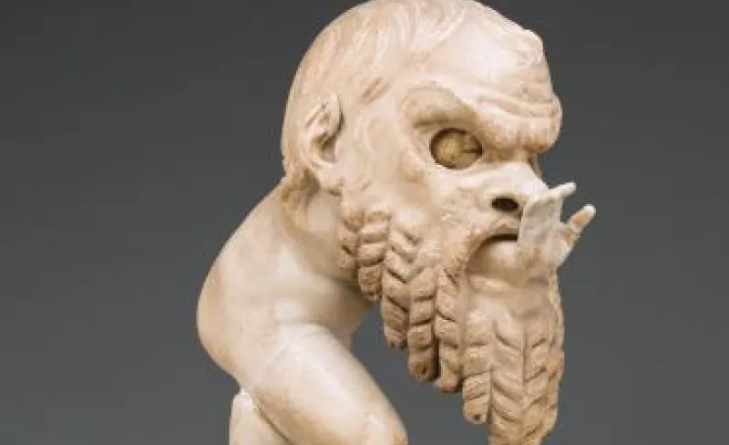 Imagen parcial de una escultura romana antigua en mármol blanco. Representa a un bebé regordete, un pequeño sátiro, que jugando se ha metido dentro de una gran máscara de teatro: se ve la parte superior de su cara por el hueco para los ojos, y asoma su manita extendida por la boca de la máscara.