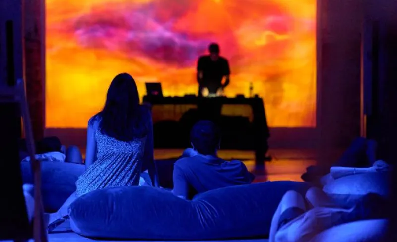 Un DJ con fondo en tonos naranjas actúa para un público sentado en el suelo