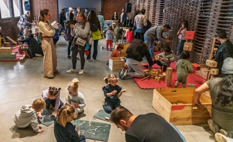 Madres padres y niños y niñas juegan con artilugios de madera en un espacio diáfano