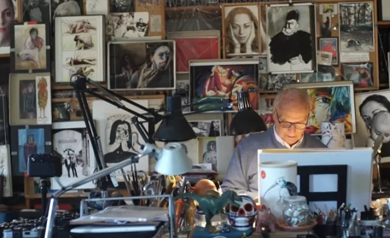 Fotografía del director de cine Carlos Saura trabajando frente a un atril con una pared de fondo llena de cuadros