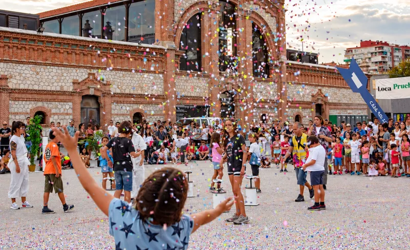 Una niña arroja confeti al cielo en una explanada junto a otros niños. De fondo un edificio del Matadero Madrid