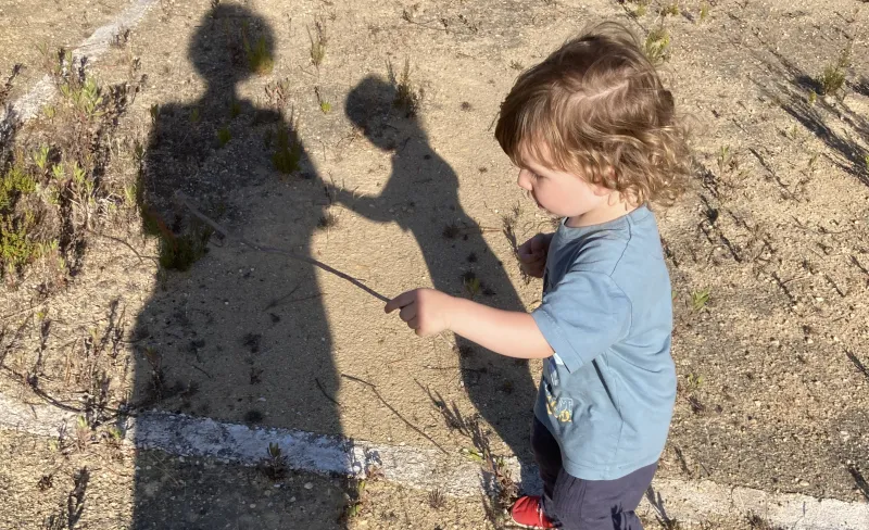 Un niño de 2 años juega con un palo observado por su madre. las sombras de ambos se proyectan en el suelo