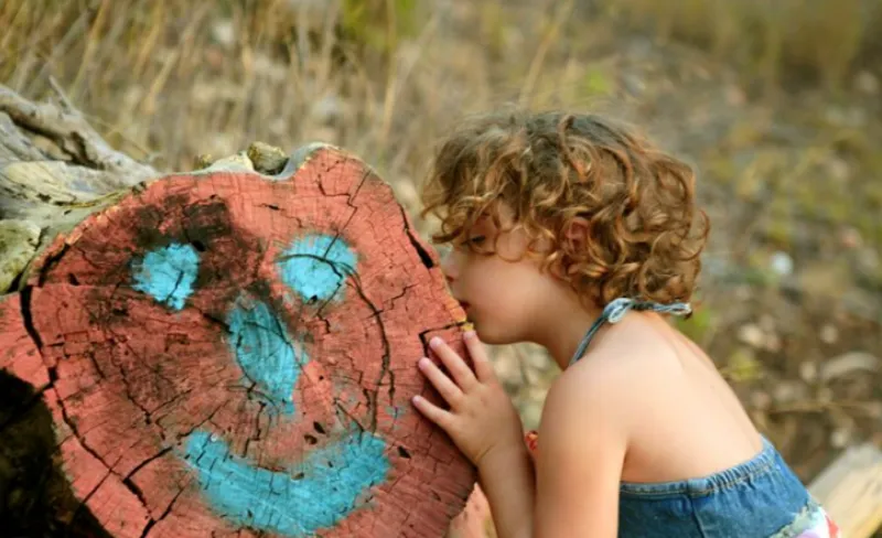una niña besa el tronco de un árbol que ha sido pintado ilustrando una cara en color azul