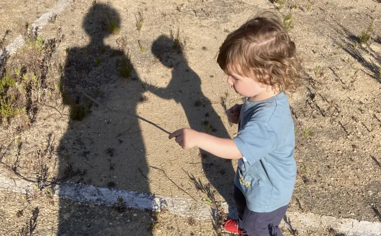 Un niño de 2 años juega con un palo observado por su madre. las sombras de ambos se proyectan en el suelo