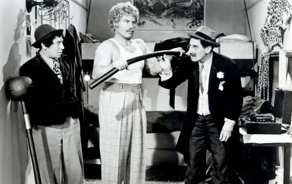 Hombre con bigote y sobrero, Groucho Marx, mira incrédulo a un forzudo que dobla una barra