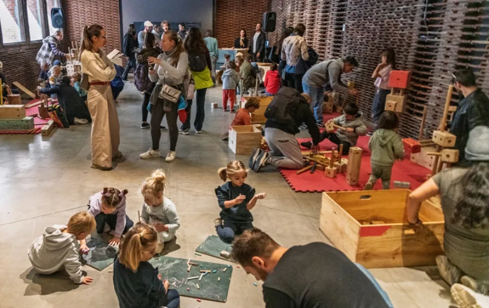 Madres padres y niños y niñas juegan con artilugios de madera en un espacio diáfano