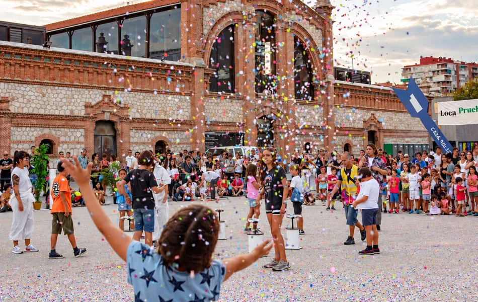 Una niña arroja confeti al cielo en una explanada junto a otros niños. De fondo un edificio del Matadero Madrid