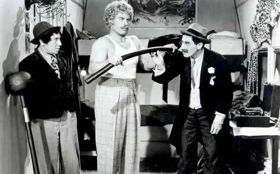 Hombre con bigote y sobrero, Groucho Marx, mira incrédulo a un forzudo que dobla una barra