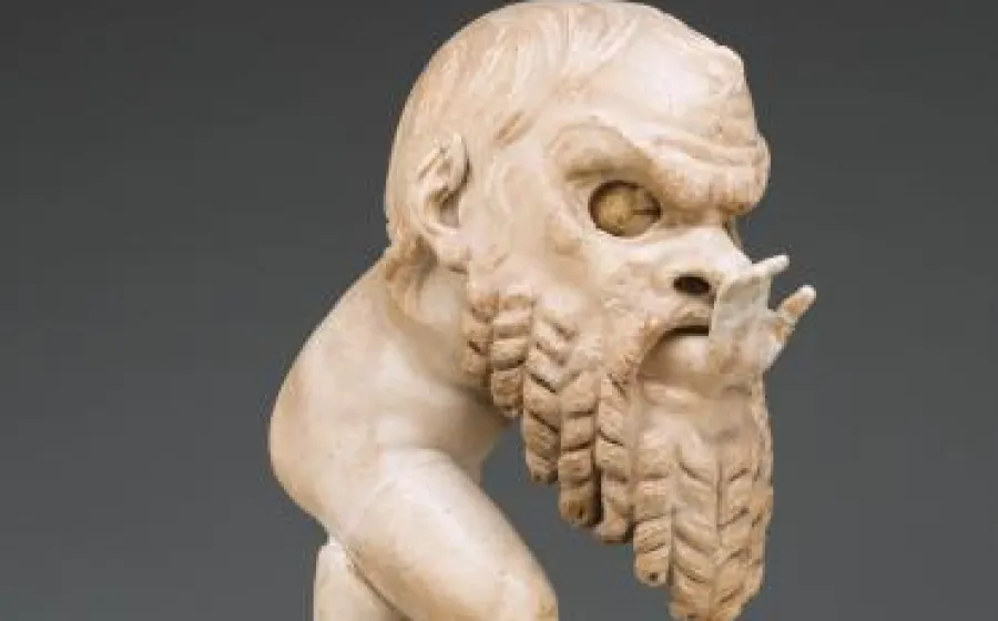 Imagen parcial de una escultura romana antigua en mármol blanco. Representa a un bebé regordete, un pequeño sátiro, que jugando se ha metido dentro de una gran máscara de teatro: se ve la parte superior de su cara por el hueco para los ojos, y asoma su manita extendida por la boca de la máscara.