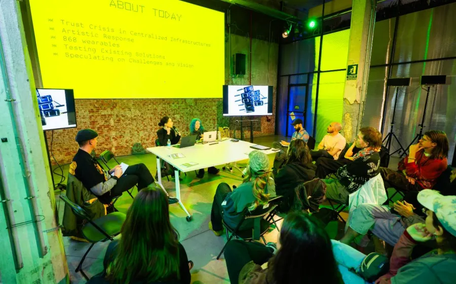 Ponentes dan una charla desde una mesa a gente con una pantalla de color amarillo detrás