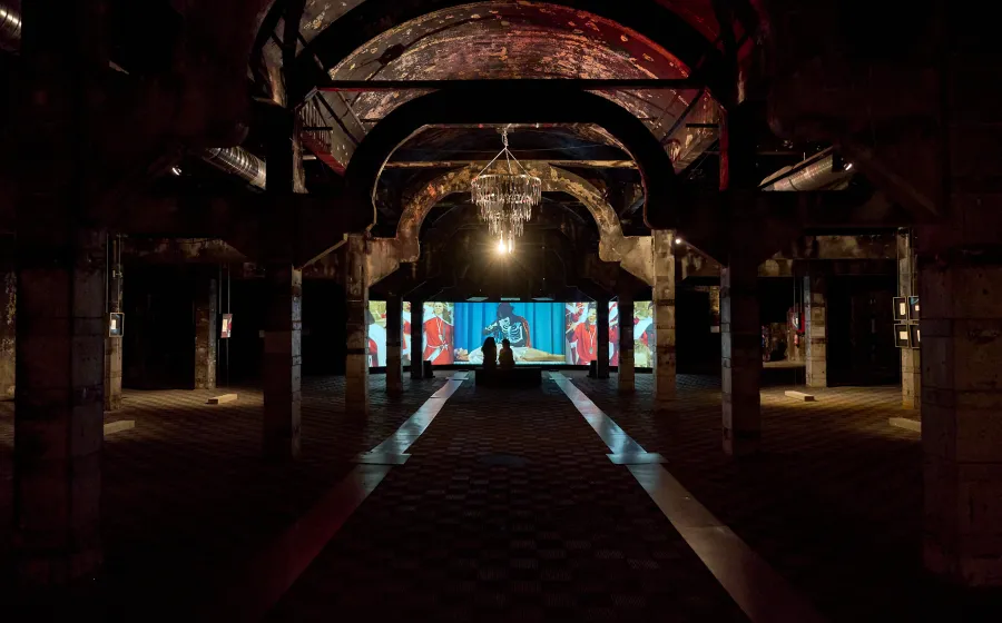 Nave industrial a oscuras con una gran lámpara iluminada y pantallas al fondo