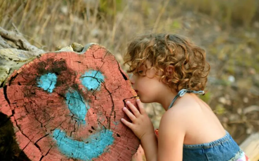 una niña besa el tronco de un árbol que ha sido pintado ilustrando una cara en color azul