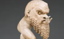Imagen parcial de una escultura romana antigua en mármol blanco. Representa a un bebé regordete, un pequeño sátiro, que jugando se ha metido dentro de una gran máscara de teatro: se ve la parte superior de su cara por el hueco para los ojos, y asoma su manita extendida por la boca de la máscara.