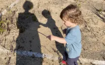 Un niño de 2 años juega con un palo observado por su madre. las sombras de ambos se proyectan en el suelo
