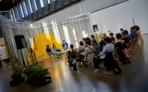 tres ponentes sentadas hablan entre sí ante una serie de asistentes con un telón amarillo y blanco de fondo