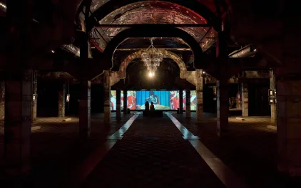 Nave industrial a oscuras con una gran lámpara iluminada y pantallas al fondo